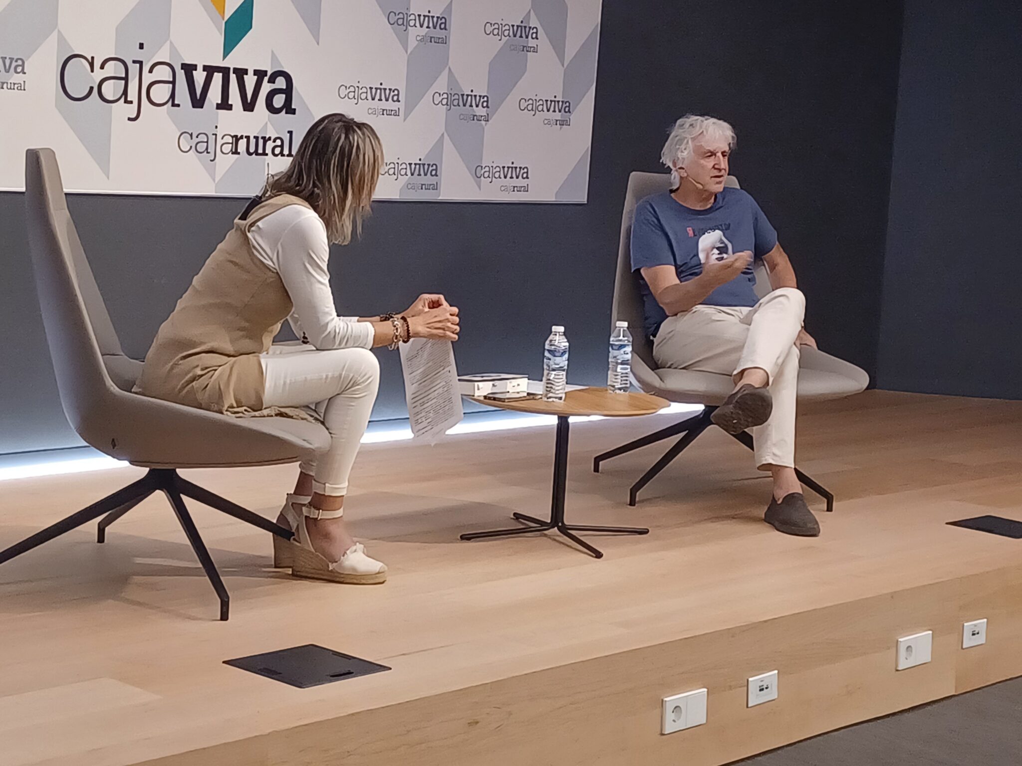 Juan Luis Arsuaga sentado en una silla en el evento Lunes por fin de Fundación Caja Rural junto con la moderadora del evento.